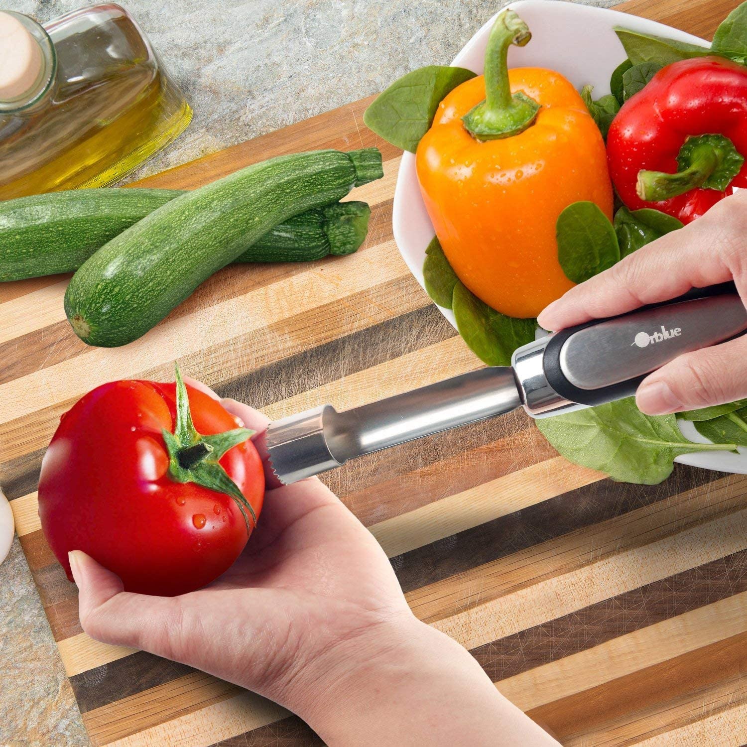 A pair of hands is using an apple corer to core a tomato. Zucchinis and capsicums are nearby on a wooden chopping board.