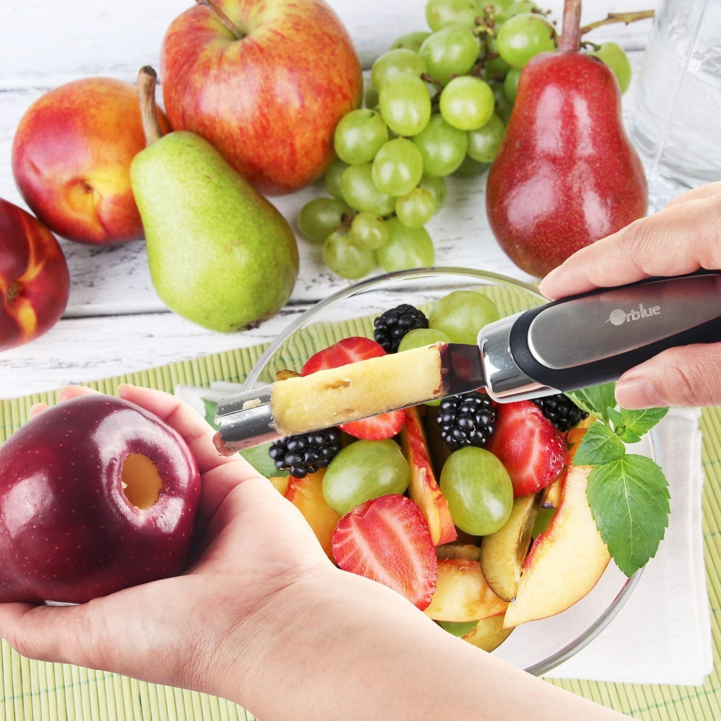 A pair of hands has just used an apple corer to core a red apple. There is also a bowl of fruit salad and various other fruits nearby.