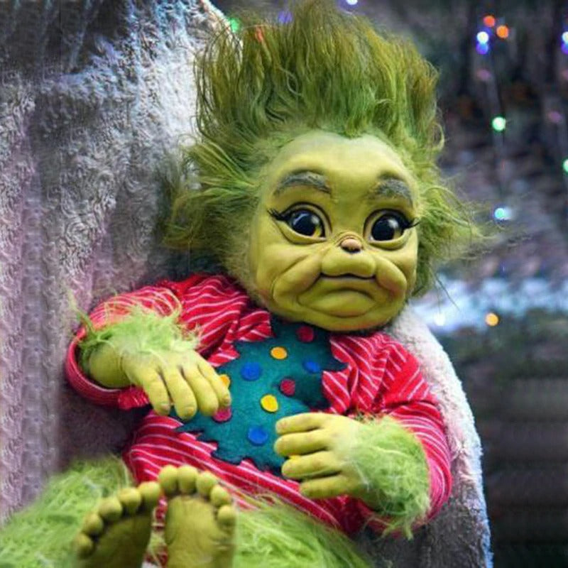 A baby grinch doll made from rubber featuring green skin and green hair wearing a Christmas costume cradling in a persons arm.
