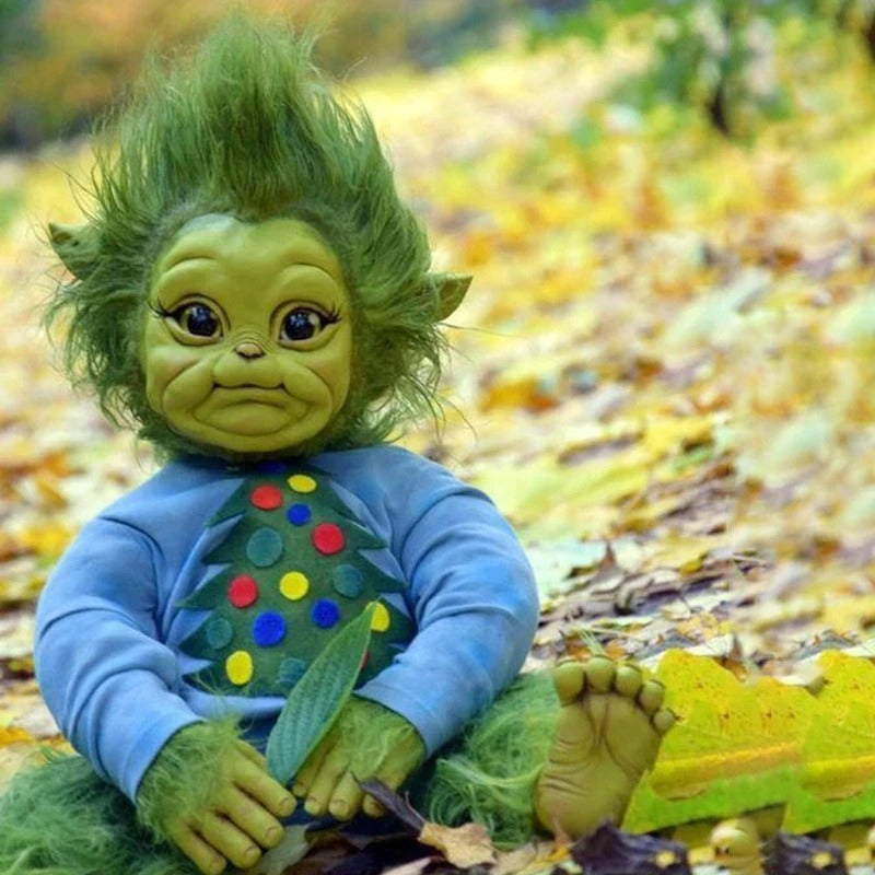 A baby grinch doll made from rubber featuring green skin and green hair wearing a blue Christmas themed t-shirt sitting on the ground surrounded by leaves.