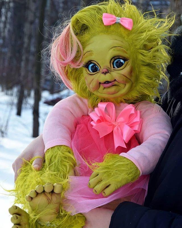 A baby grinch girl doll made from rubber featuring green skin and green hair wearing a pink costume cradling in a persons arm.