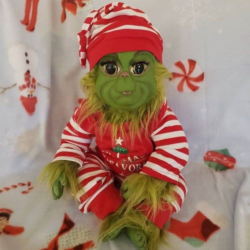 A baby grinch doll made from rubber featuring green skin and green hair wearing a Christmas costume and hat sitting on a Christmas themed blanket.