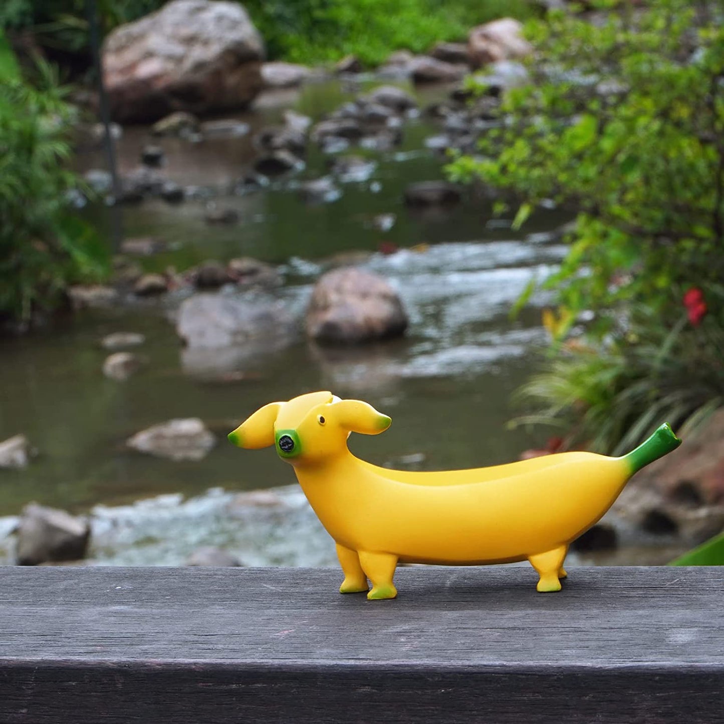 A banana dog sitting on a wooden railing of a bridge with a river behind him. A banana dog is a garden decoration which is a banana shaped dog.
