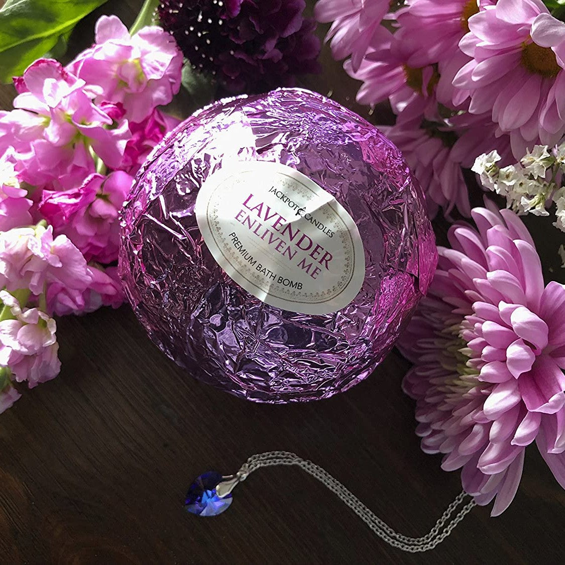 A bath bomb wrapped in purple foil with a label that says, 'lavender.' There is a blue heart shaped necklace next to the bath bomb. There are purple flowers surrounding the bath bomb and necklace.