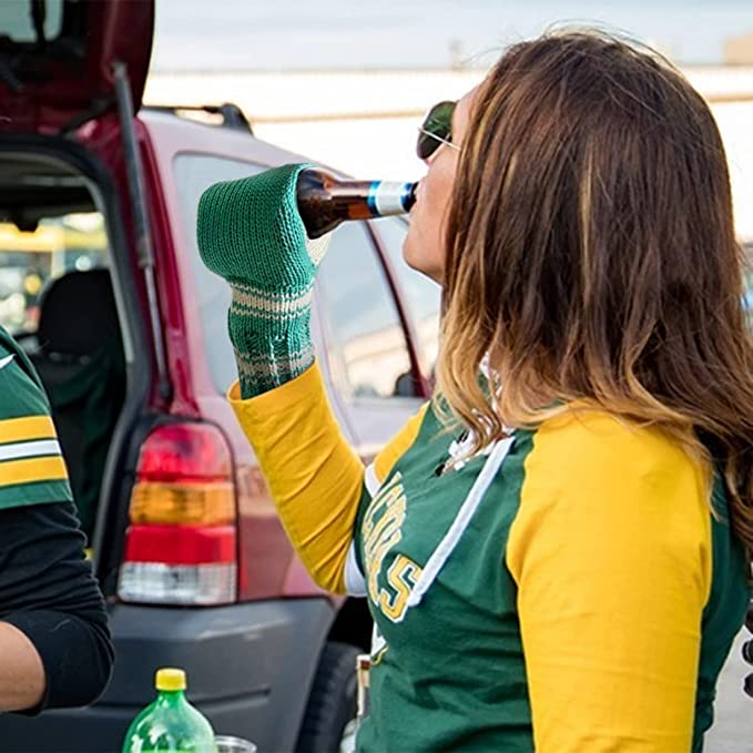 A woman is drinking a bottle of beer while wearing a green beer mitt on her hand.