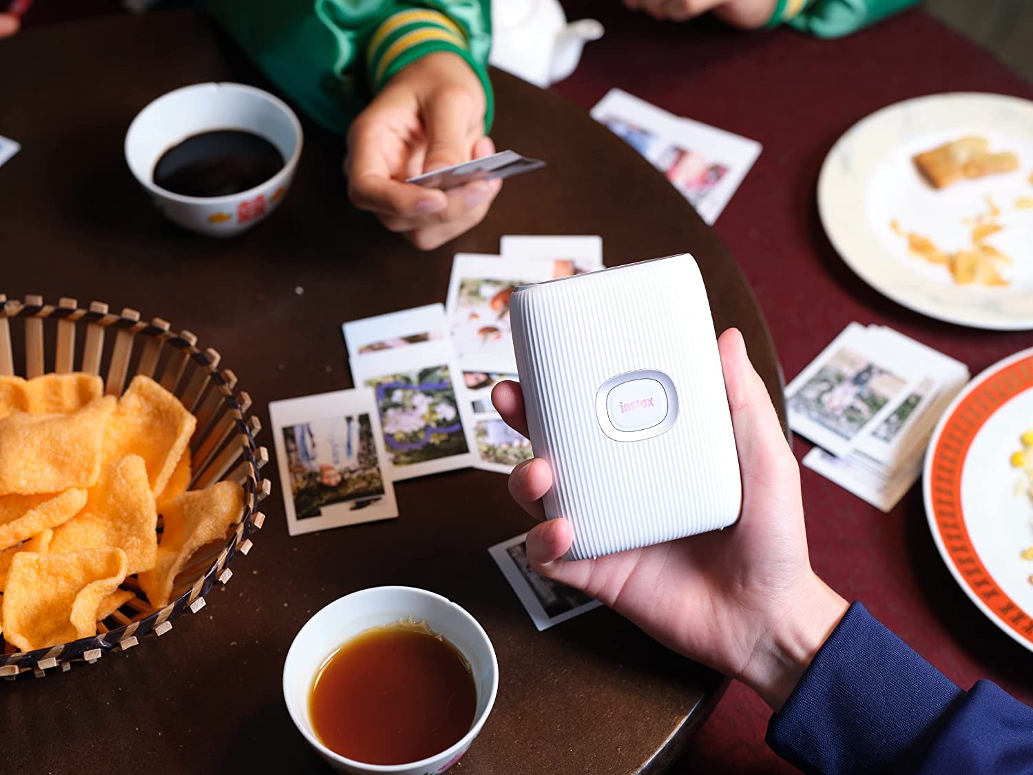 A hand is holding a Bluetooth smartphone printer while at a table. Another hand is holding a photo while other photos are scattered around on the table.
