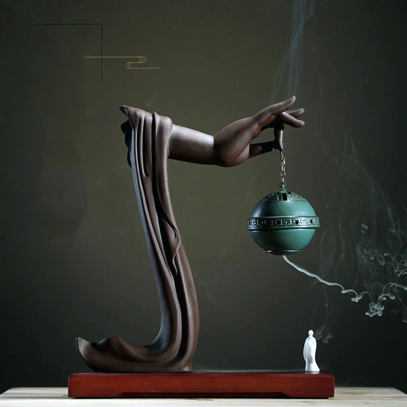 A backflow incense holder. There is a copper colored Buddha hand holding an ornate incense holder attached to a chain. There is incense smoke coming out of the holder. Beneath the holder is a white figure waiting to be engulfed by the soothing fog of incense.