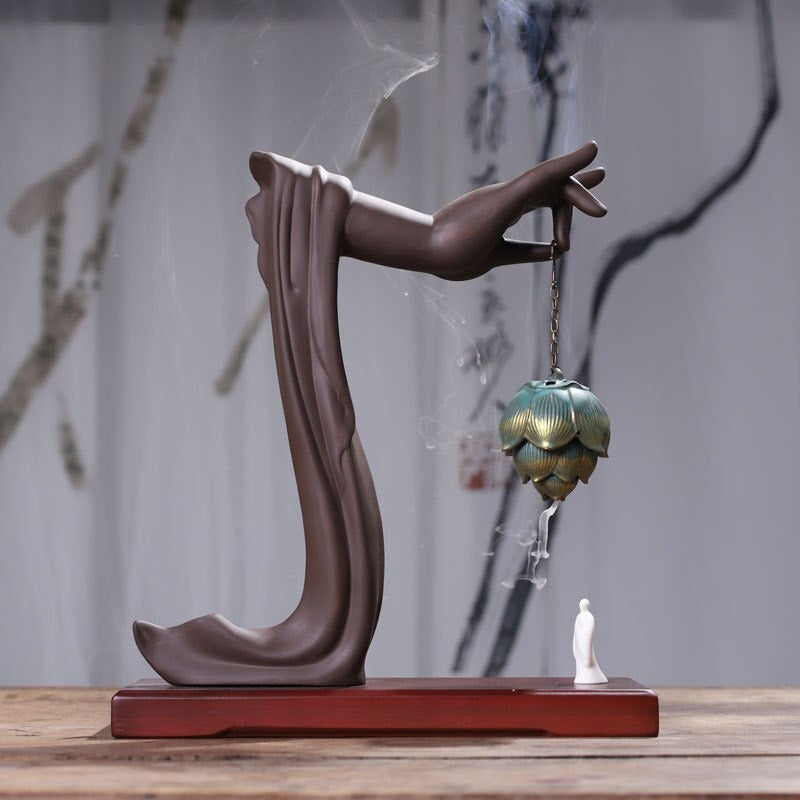 A backflow incense holder on a reddish brown base. There is a brown colored Buddha hand holding an acorn shaped incense holder attached to a chain. There is incense smoke coming out of the holder. Beneath the holder is a white figure.