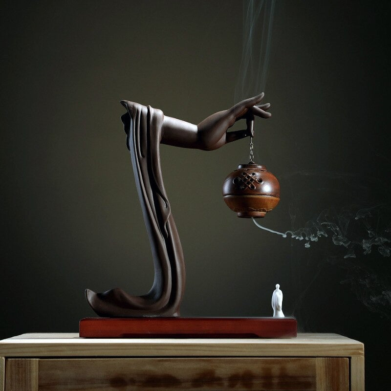 A backflow incense holder on a reddish brown base and dark green background. There is a brown colored Buddha hand holding a red sphere-shaped incense holder attached to a brown chain. There is incense smoke bellowing out of the holder. Beneath the holder is a white figure.