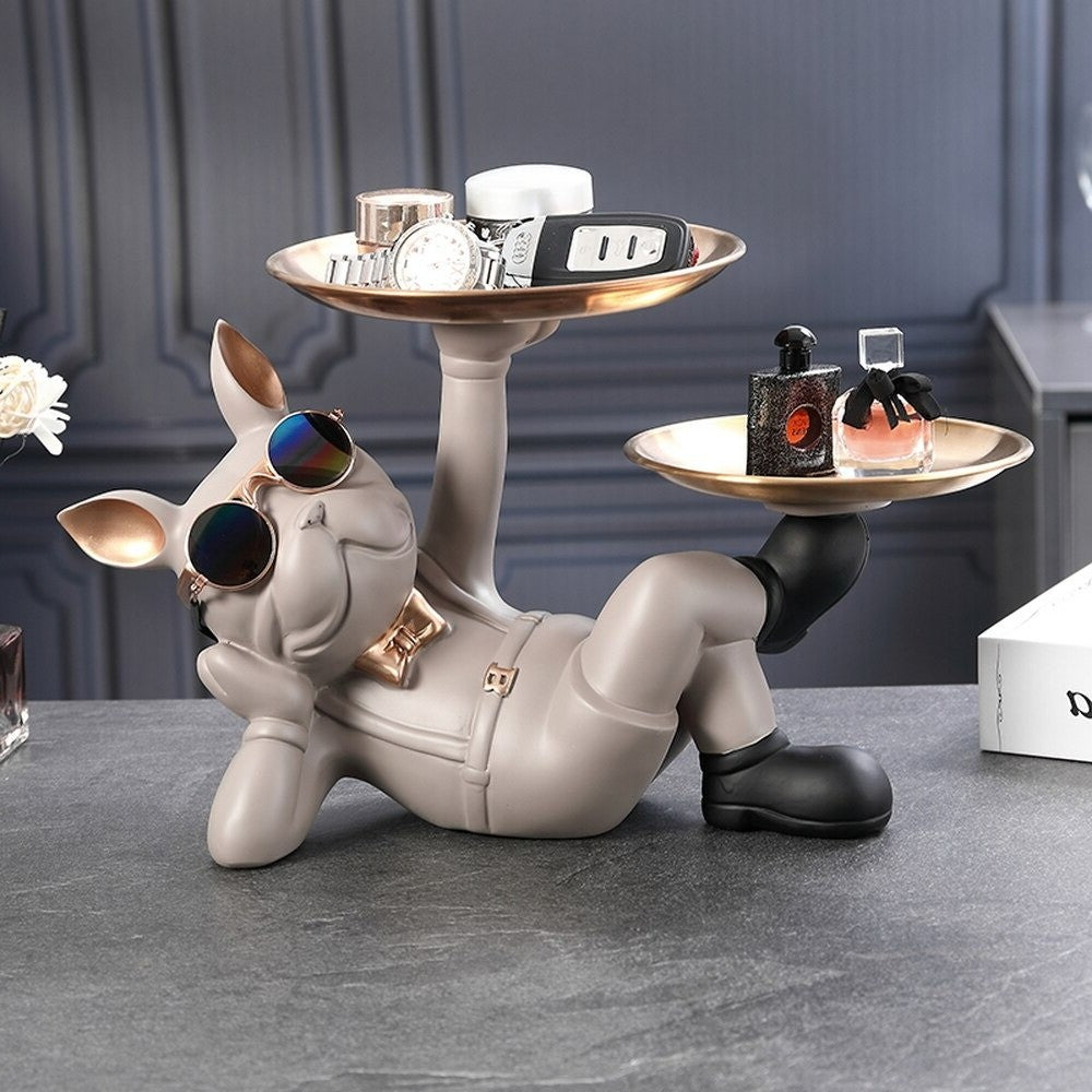 A resin bulldog serving tray. The bulldog is gray and reclined in a relaxed pose holding two serving trays, one tray is held by his hand and the other is held by his foot. There are knick knacks on both trays. He is wearing a pair of cool sunglasses and a cute gold bow tie.