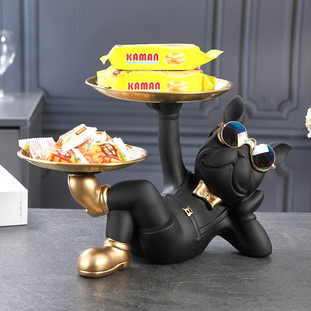 A resin bulldog serving tray. The bulldog is black and reclined in a relaxed pose holding two serving trays, one tray is held by his hand and the other is held by his foot. There are knick knacks on both trays. He is wearing a pair of cool sunglasses and a cute gold bow tie.