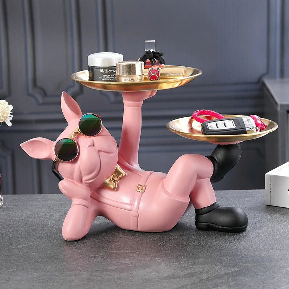 A resin bulldog serving tray. The bulldog is pink and reclined in a relaxed pose holding two serving trays, one tray is held by his hand and the other is held by his foot. There are knick knacks on both trays. He is wearing a pair of cool sunglasses and a cute gold bow tie.