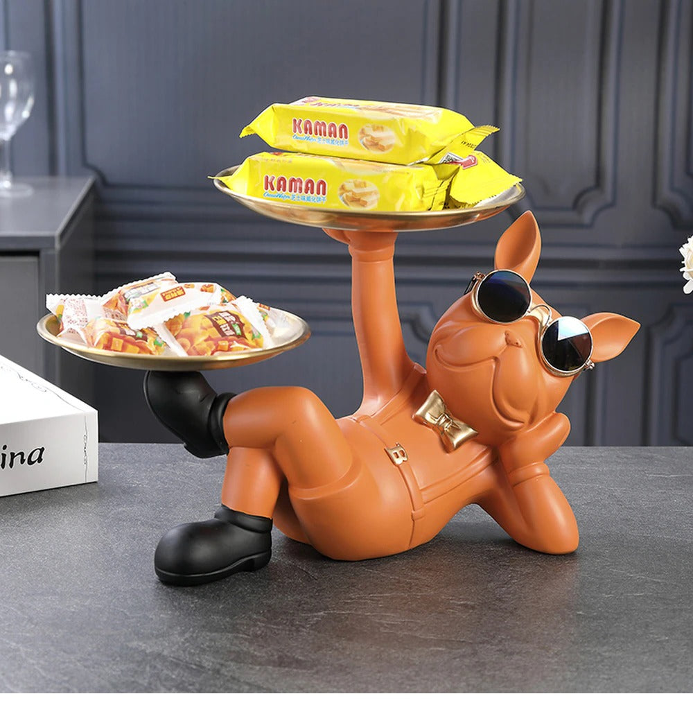 A resin bulldog serving tray. The bulldog is orange and reclined in a relaxed pose holding two serving trays, one tray is held by his hand and the other is held by his foot. There are knick knacks on both trays. He is wearing a pair of cool sunglasses and a cute gold bow tie.