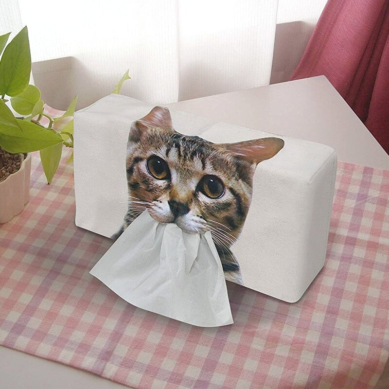 A rectangle tissue box holder which has a cats face printed on the front and has tissues coming out of its mouth.