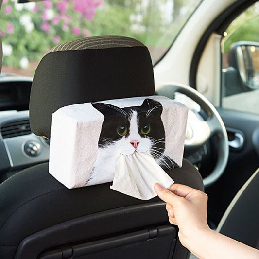 A cat face tissue box holder on the back of a car seat. There is a cats face printed on the front and a hand pulling a tissue out of its mouth.