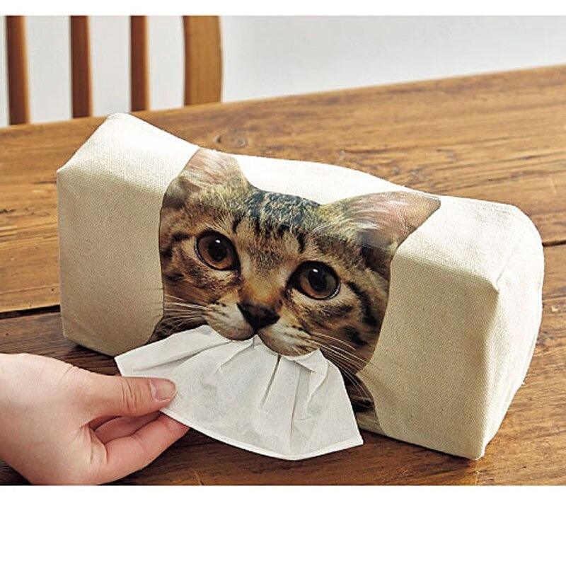 A hand is pulling a white tissue holder which has a cats face printed on the front of it with tissues dispensed from the mouth.