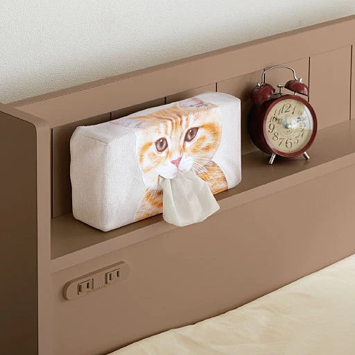 A cat face tissue box holder which has a cats face printed on the front of it. The holder is sitting on a shelf within a bedhead