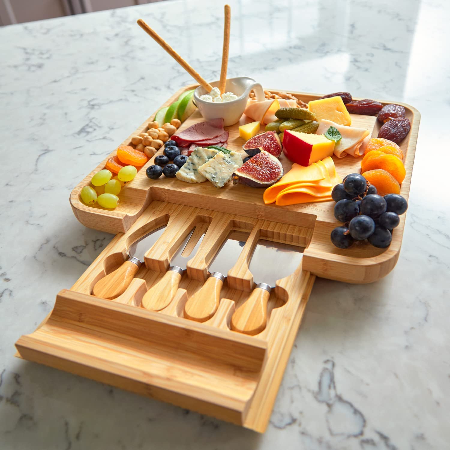 heese board and knife set with slide-out cutlery drawer. On the board are various different foods including, cheeses and fruits.
