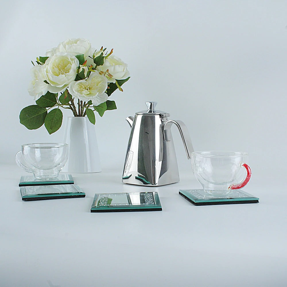 A set of four silver coasters filled with tiny crystals, 2 transparent cups sit on 2 of the coasters and a silver kettle is nearby plus a bunch of white flowers.