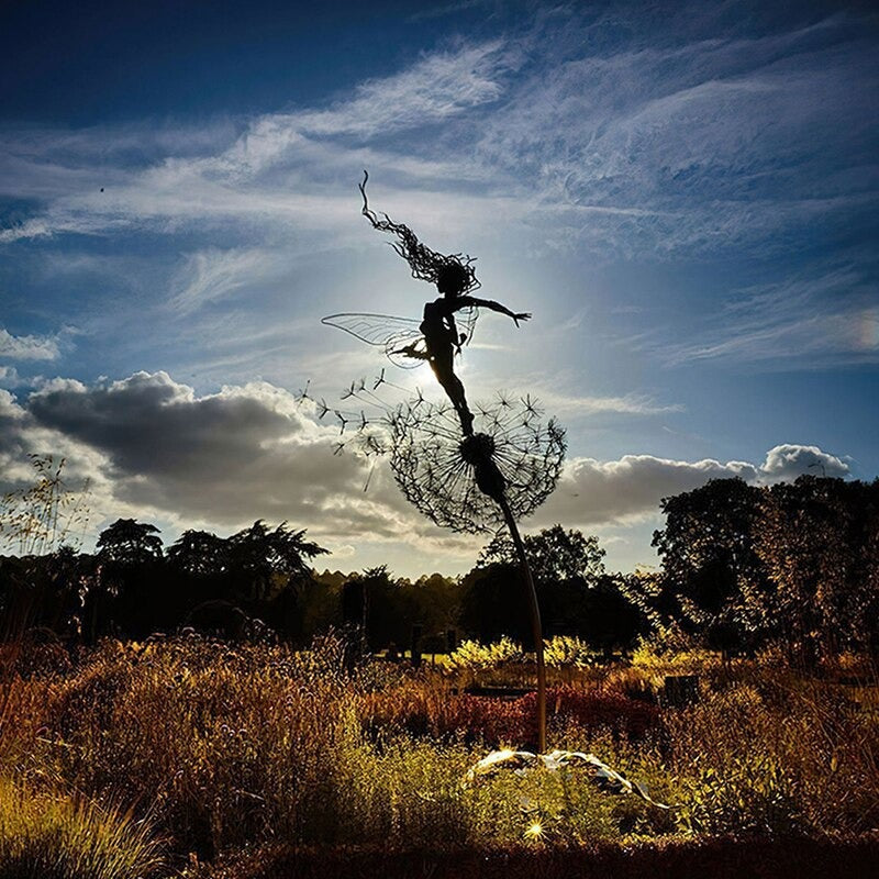 A beautiful metal fairy garden sculpture. The sculpture is of a fairy with wings leaping into the air while standing on top of a dandelion.