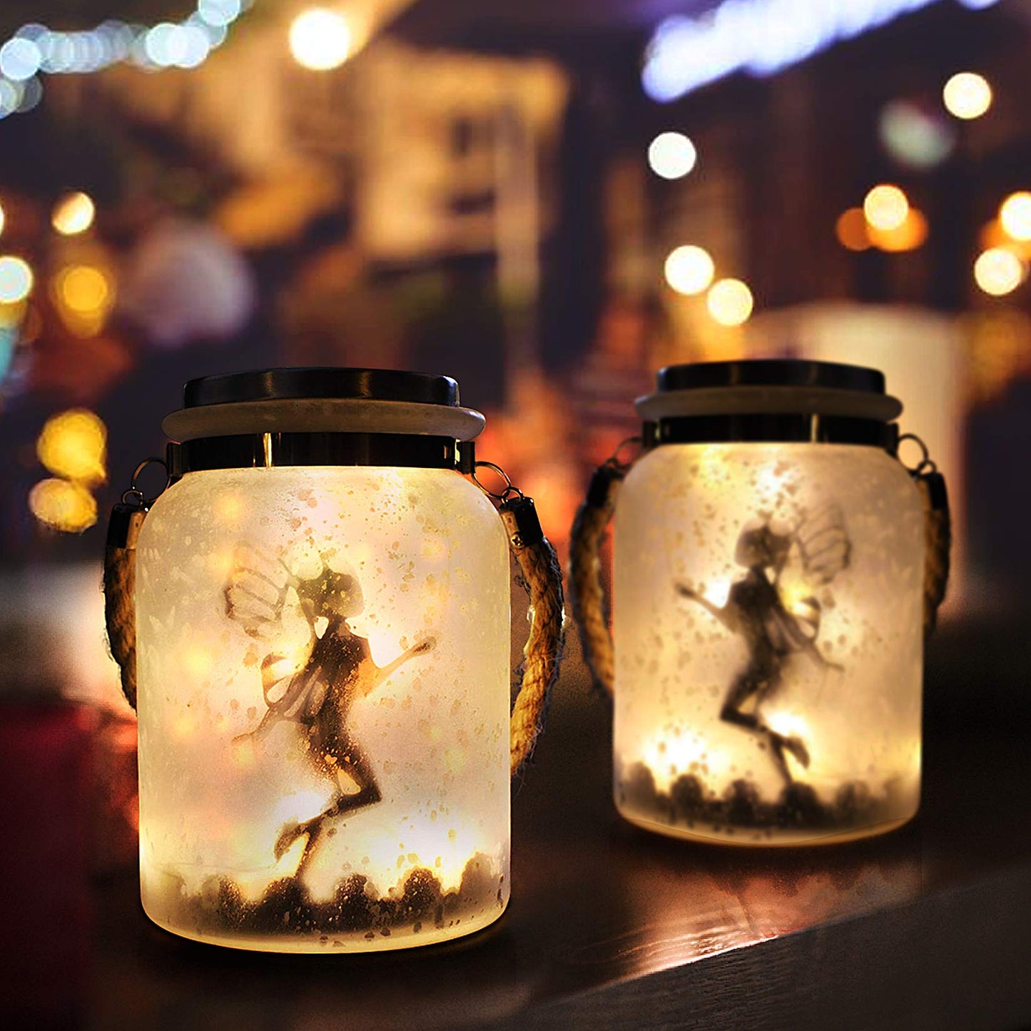 Two solar mason jars lit up with the silhouette of 2 fairies inside set to a backdrop at night.