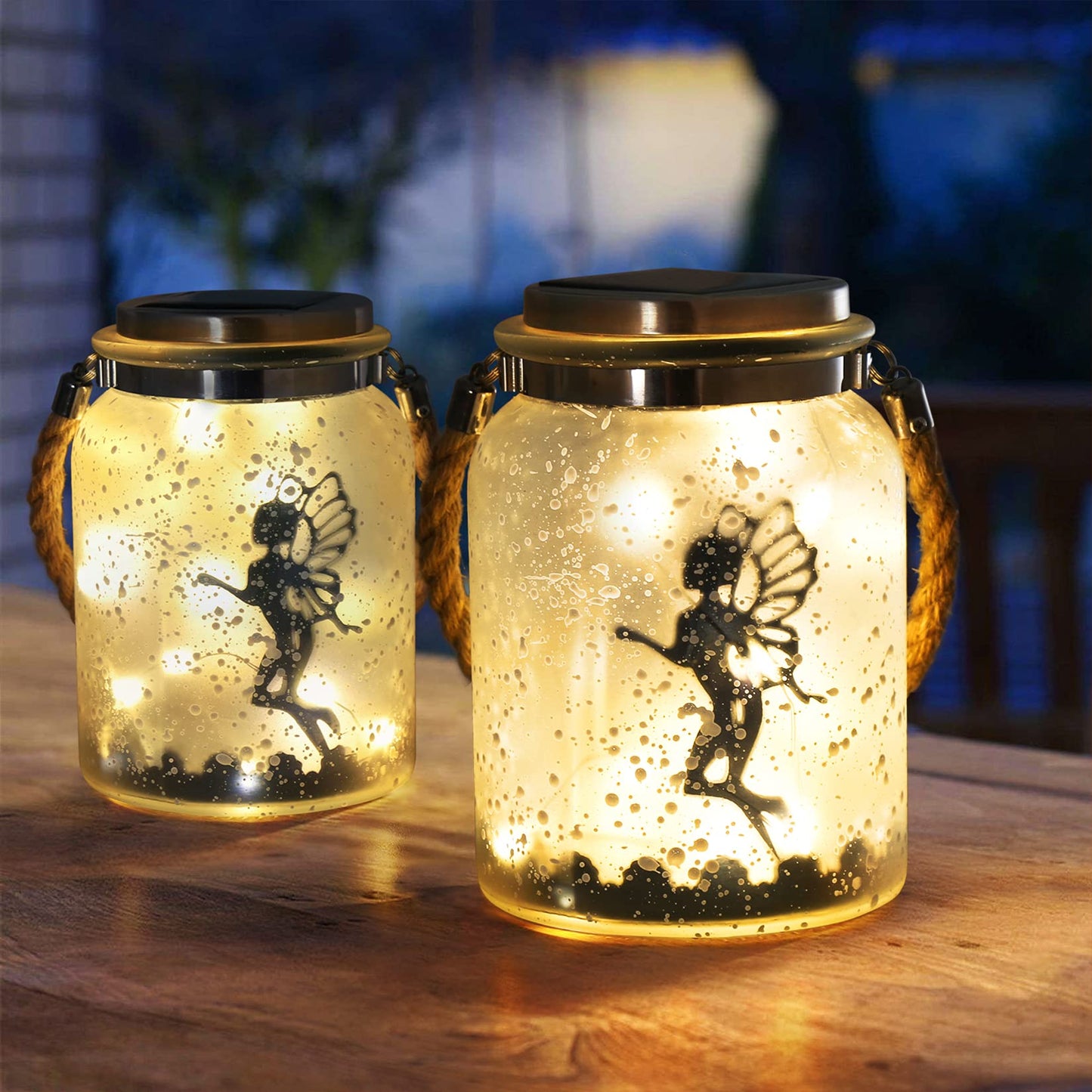 Two solar mason jars lit up with the silhouette of 2 fairies inside set to a backdrop at night.