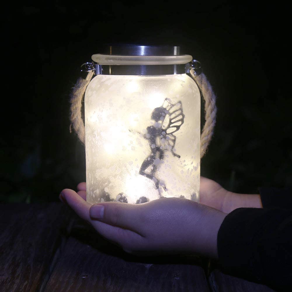 A pair of hands holding a solar mason jar lit up with the silhouette of 2 fairies inside at night.