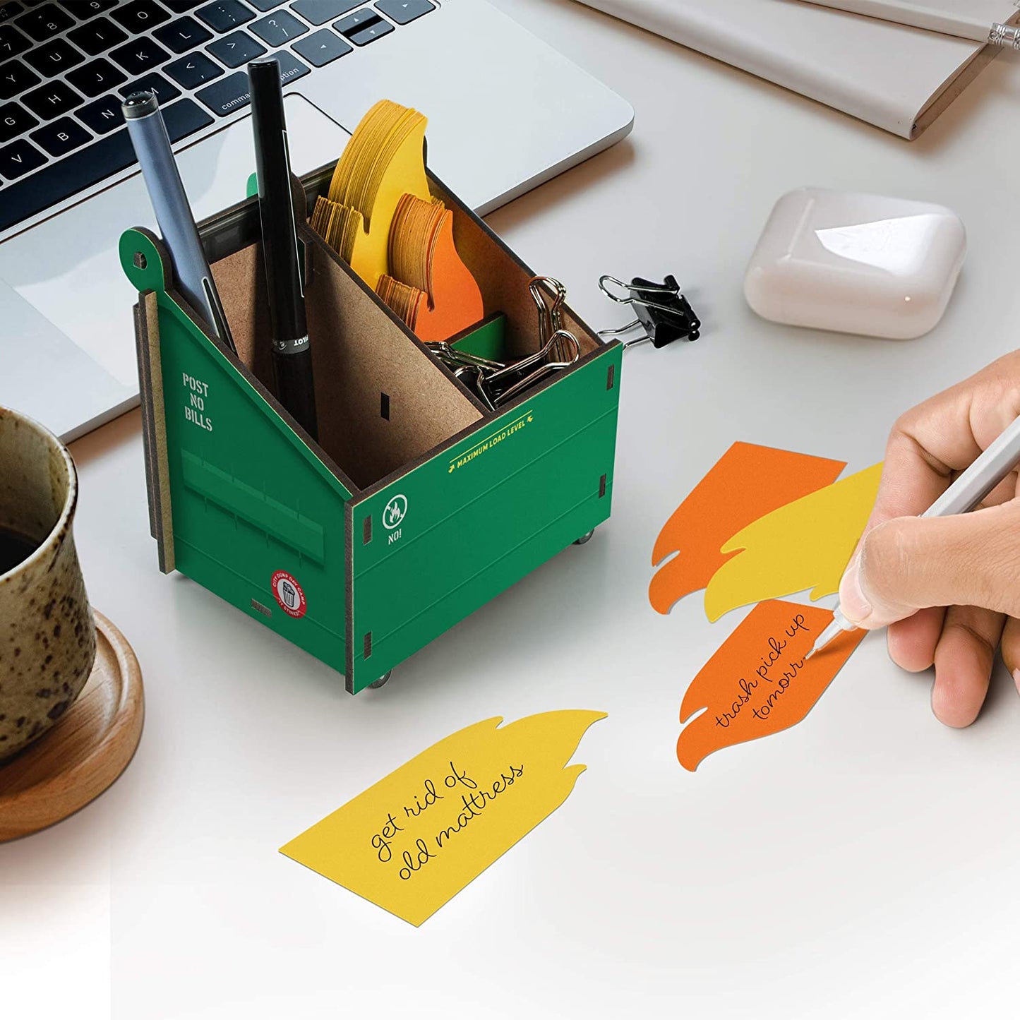 A pencil and note holder in the shape of a dumpster fire is on a desk. The note cards look like flames and a hand is writing on one of the notes.
