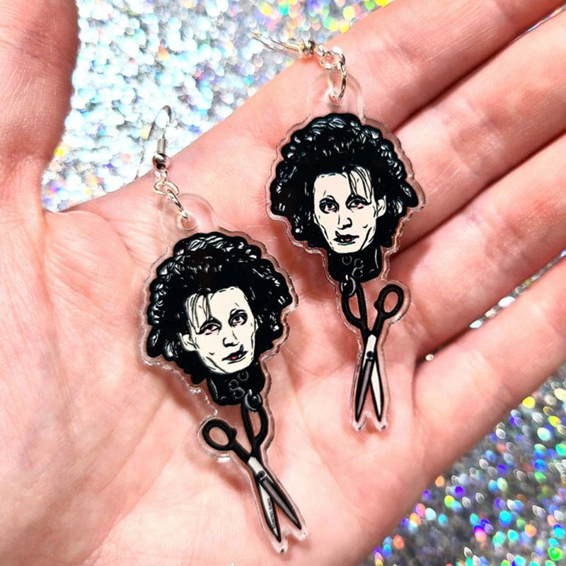 A pair of earrings inspired by the 90's movie Edward Scissorhands. They feature Edward Scissorhands head with a pair of scissors handing off the end. These are being held in a persons palm.