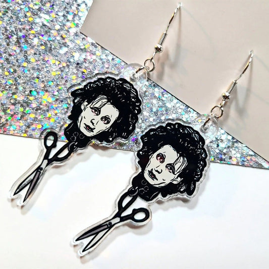 A pair of earrrings inspired by the 90's movie Edward Scissorhands. They feature Edward Scissorhands head with a pair of scissors handing off the end. These are resting on a white and silver glitter background.