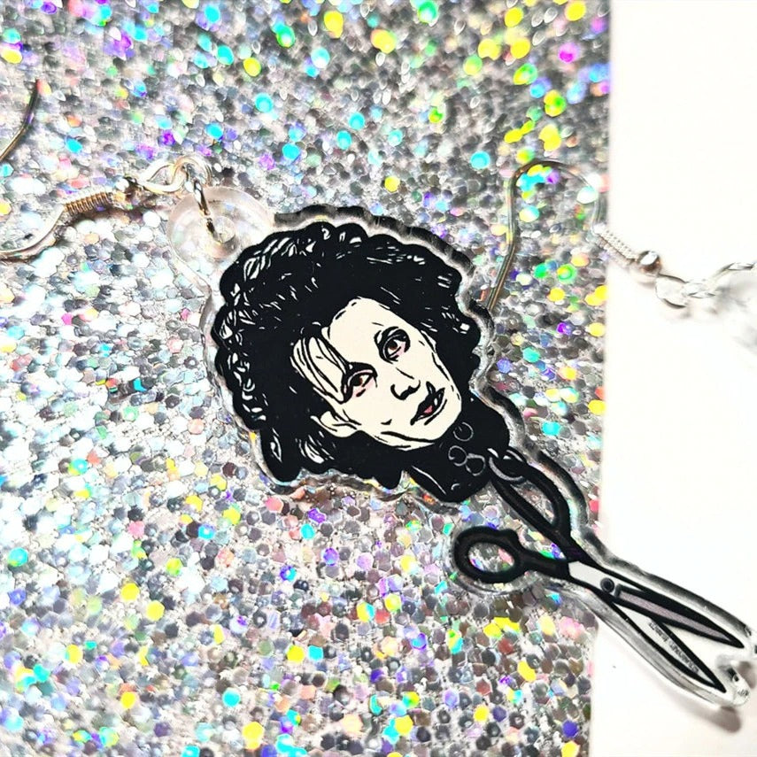 Close up view of one earrring inspired by the 90's movie Edward Scissorhands. It features Edward Scissorhands head with a pair of scissors handing off the end. These are resting on a white and silver glitter background.