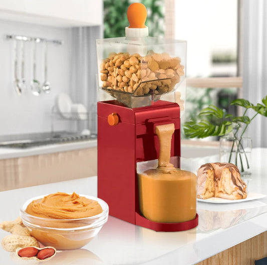 A red electric peanut butter machine in operation with peanut butter pouring out into a container under the machine. There is a fresh bowl of peanut butter next to the machine as well as a cake.