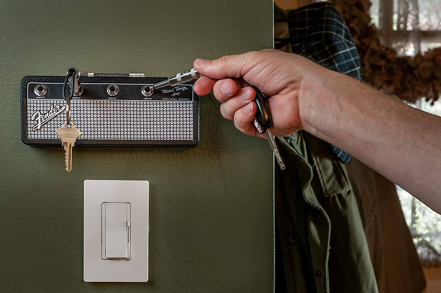 A Fender key rack holder on a green wall. A persons hand is putting one of the included keychain jacks into one of the plugs which holds the keys.