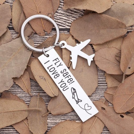 A keyring which has a tag that says fly safe, I love you. There is a silver airplane attached to the keychain as well. The whole keychain rests on brown leaves.