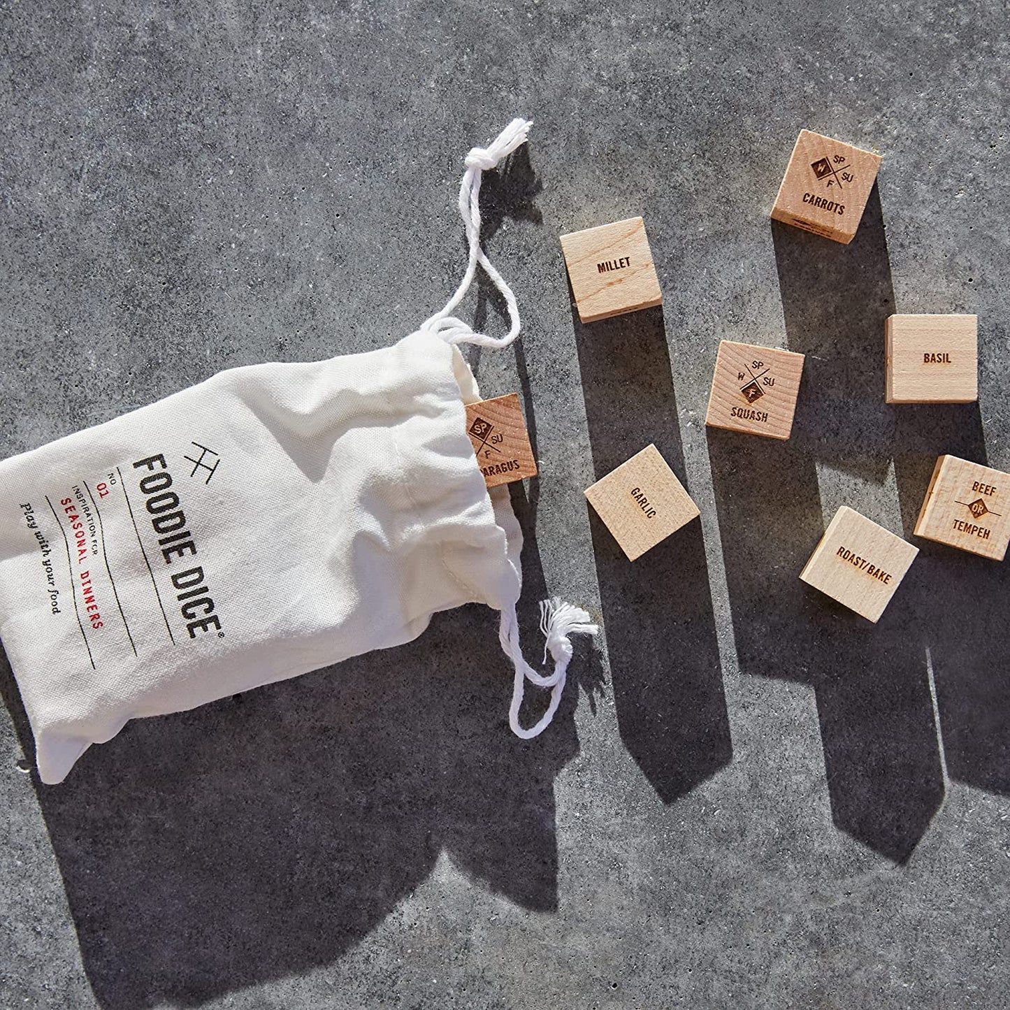 Foodie dice which is a set of dice which help a person to decide what to cook for dinner. Eight dice are scattered about with a white satchel nearly which says "Foodie Dice" on it.