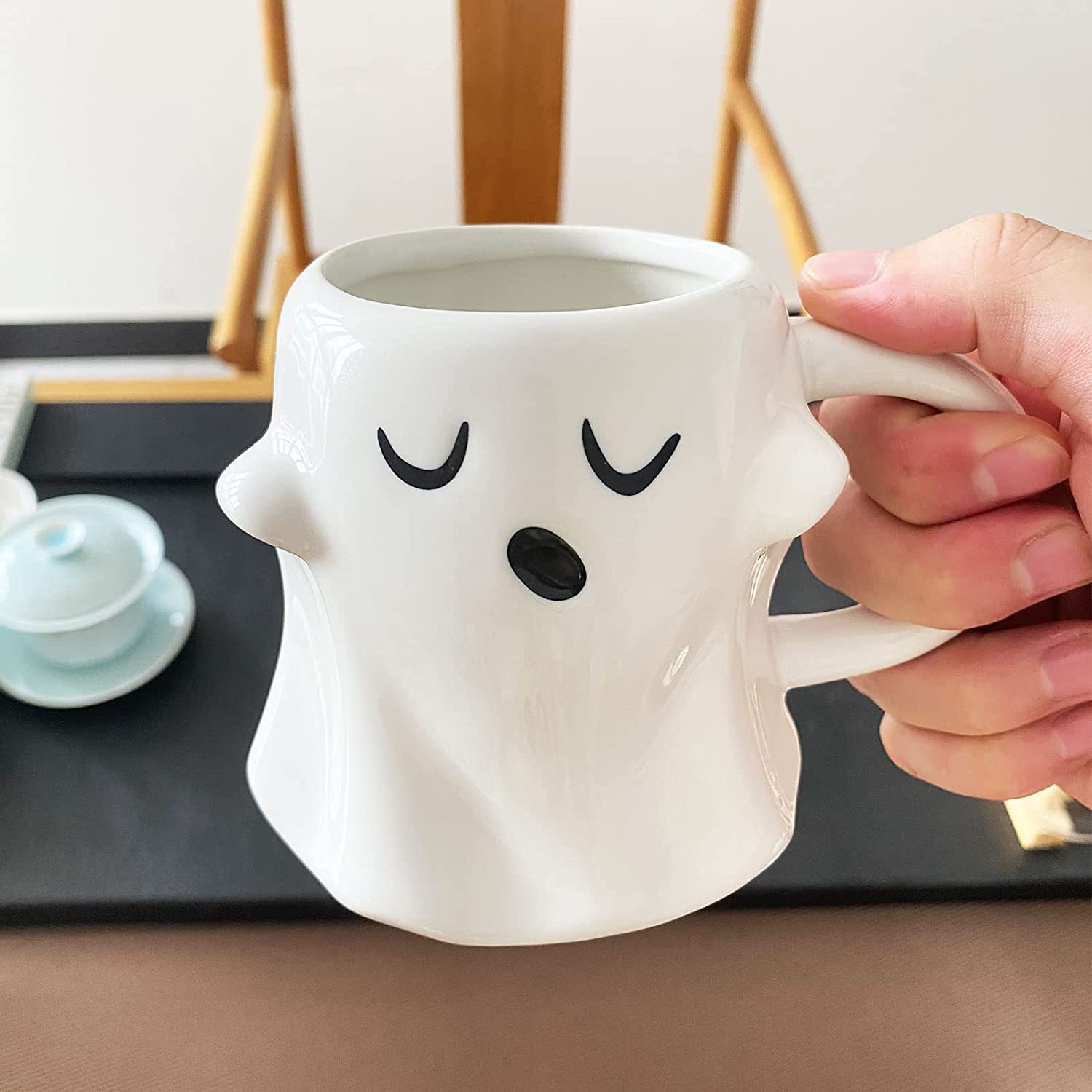 A hand holding a white ceramic ghost shaped coffee mug which features an expression as if it is yawning and waking up.