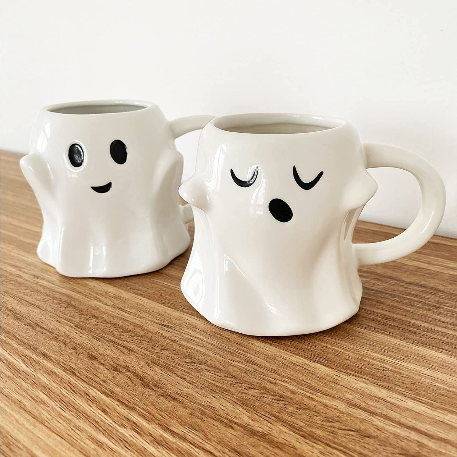 Two white ceramic ghost shaped coffee mugs on a wooden table. Both mugs have cute expressions printed on them.