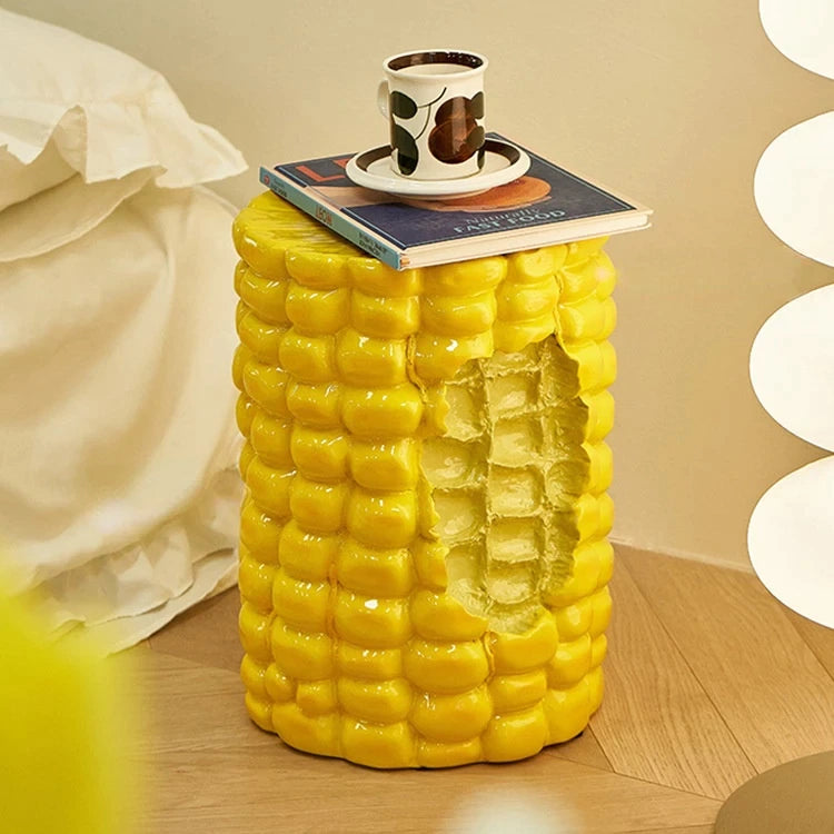 A giant cob of corn which is a footstool. A book and cup of coffee are resting on top of the corn stool.