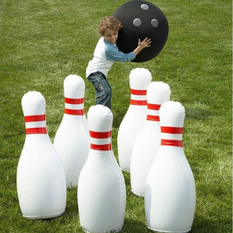 A giant inflatable outdoor bowling set. A child is holding the black inflatable ball and he is about to throw it towards the 6 pins.