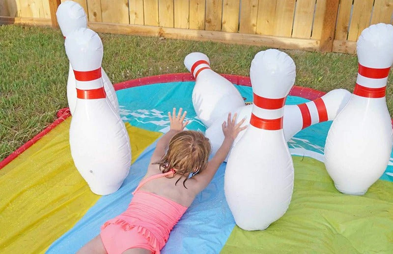 Six bowling pins set up outdoors at the end of a Slip N Slide. A child appears to have used the slid and knocked over two of the 6 pins.