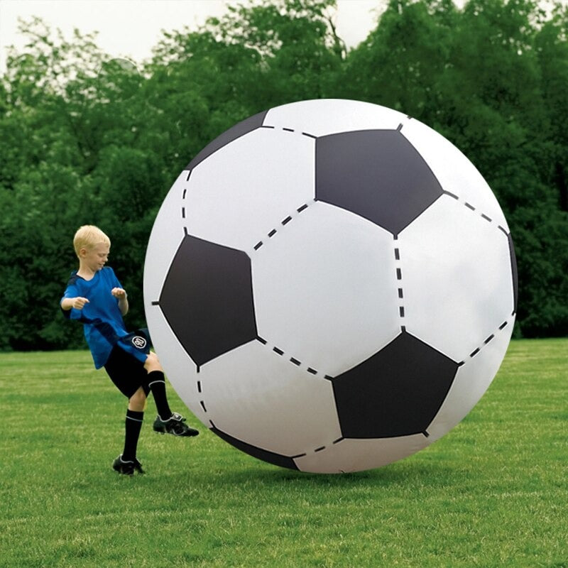 A kid playing with a giant inflatable soccer ball while in a football field. The giant ball over shadows the small kid.