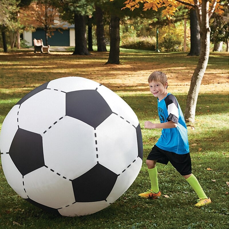A young kid playing with a giant inflatable soccer ball while in a park filled with trees. The giant soccer ball is the same height as the child.