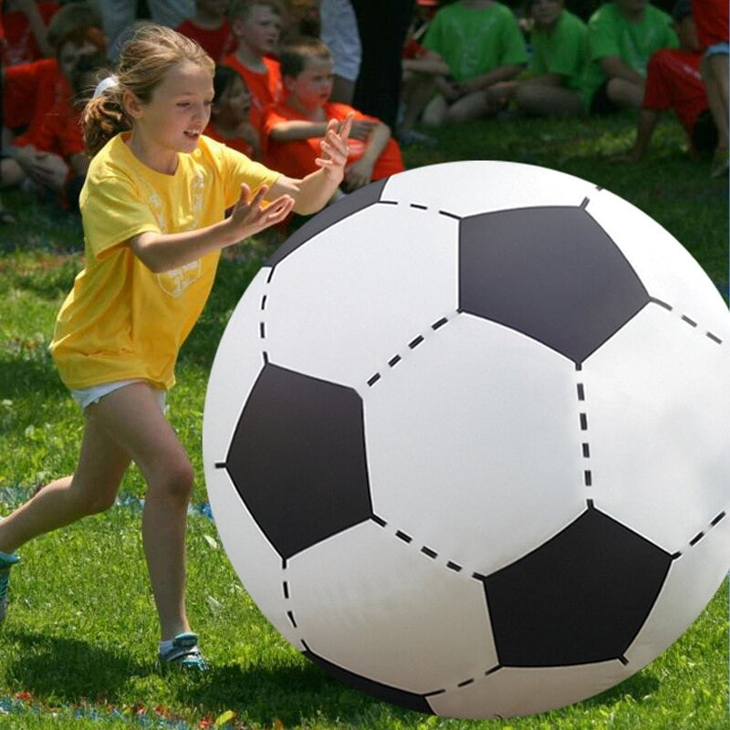A young girl pushing along a giant inflatable soccer ball. She appears to be at a school event with other children behind her. The giant soccer ball is the same height as the girl.