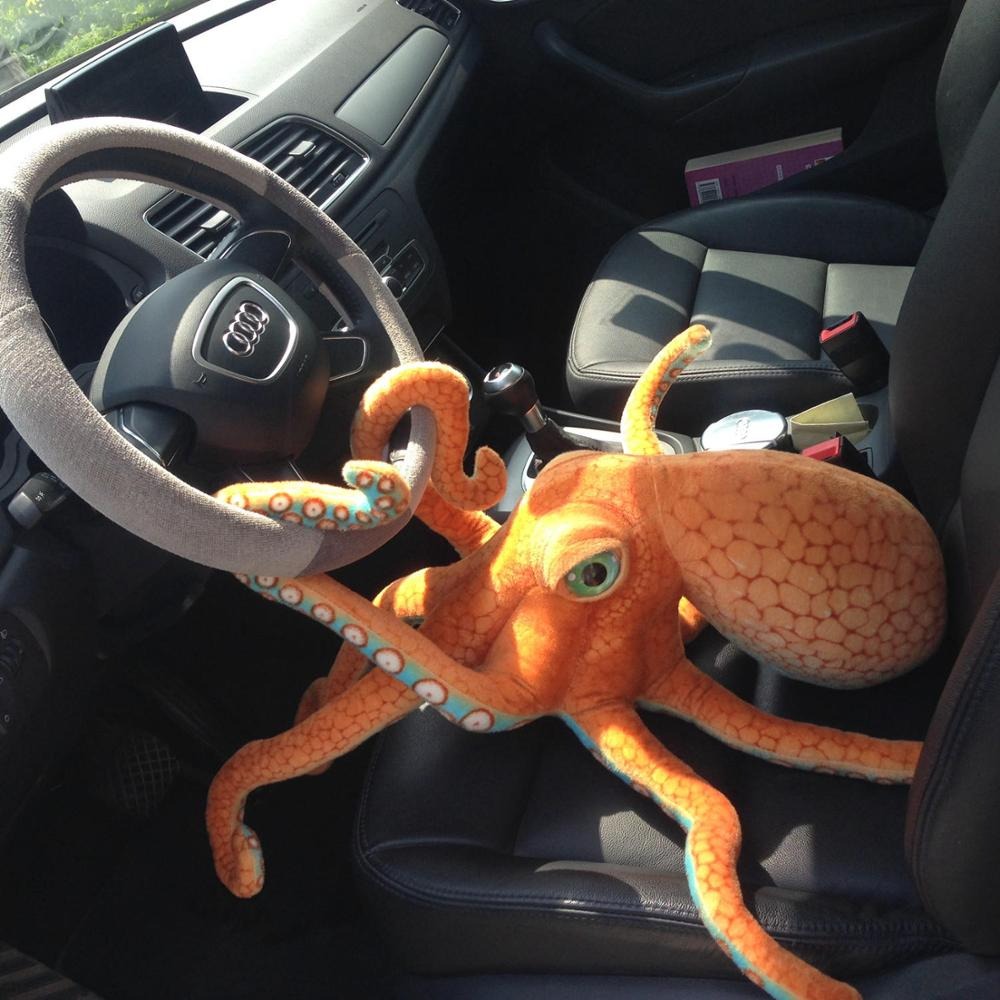 An orange plush octopus toy sitting in the drivers seat of a car with two tentacles wrapped around the steering wheel