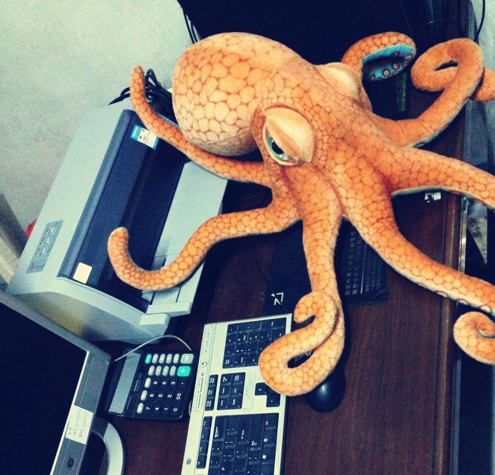 A large octopus plush toy resting on a computer keyboard and printer on a wooden desk.