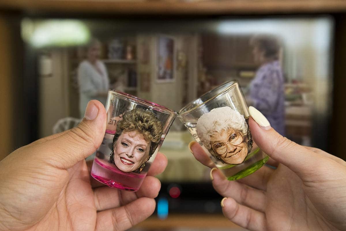Two hands are holding Golden Girls shot glasses and clinking them together. In the background is a scene from the Golden Girls tv show.