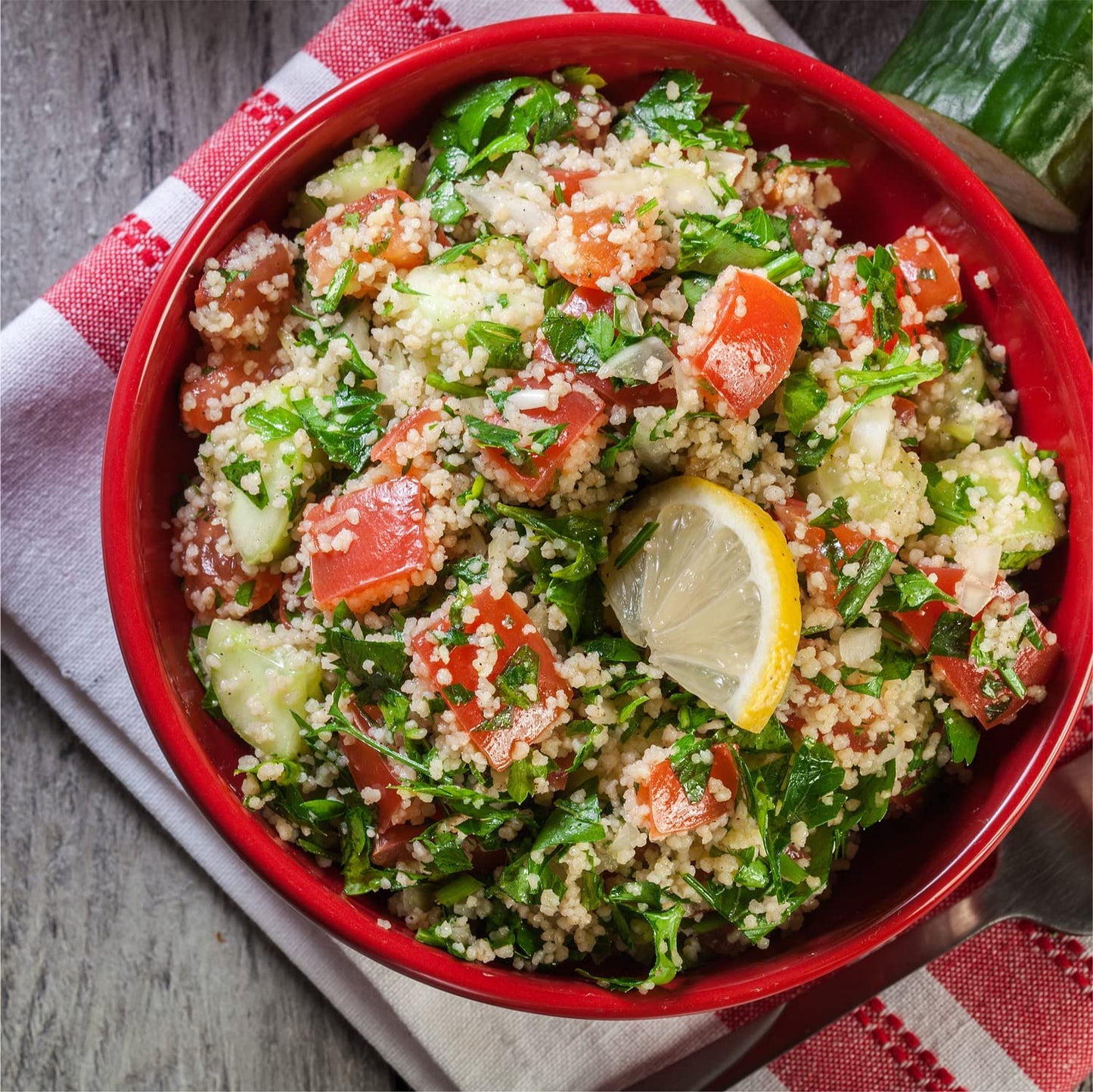 A red bowl filled with a salad made from fresh herbs, couscous and tomato with a half-lemon slice garnish.