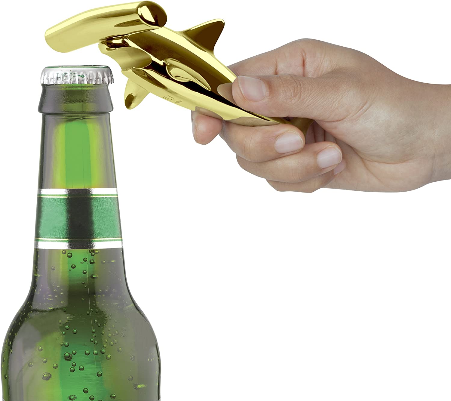 A hand using a brass colored hammerhead shark shaped bottle opener to open a bottle of beer.