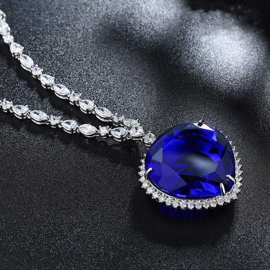 A replica of the Heart of the Ocean necklace from the Titanic movie. This is a closeup of the main pendant which shows the blue gem. Some links of the chain can also be seen. This is on a black, glittery background.