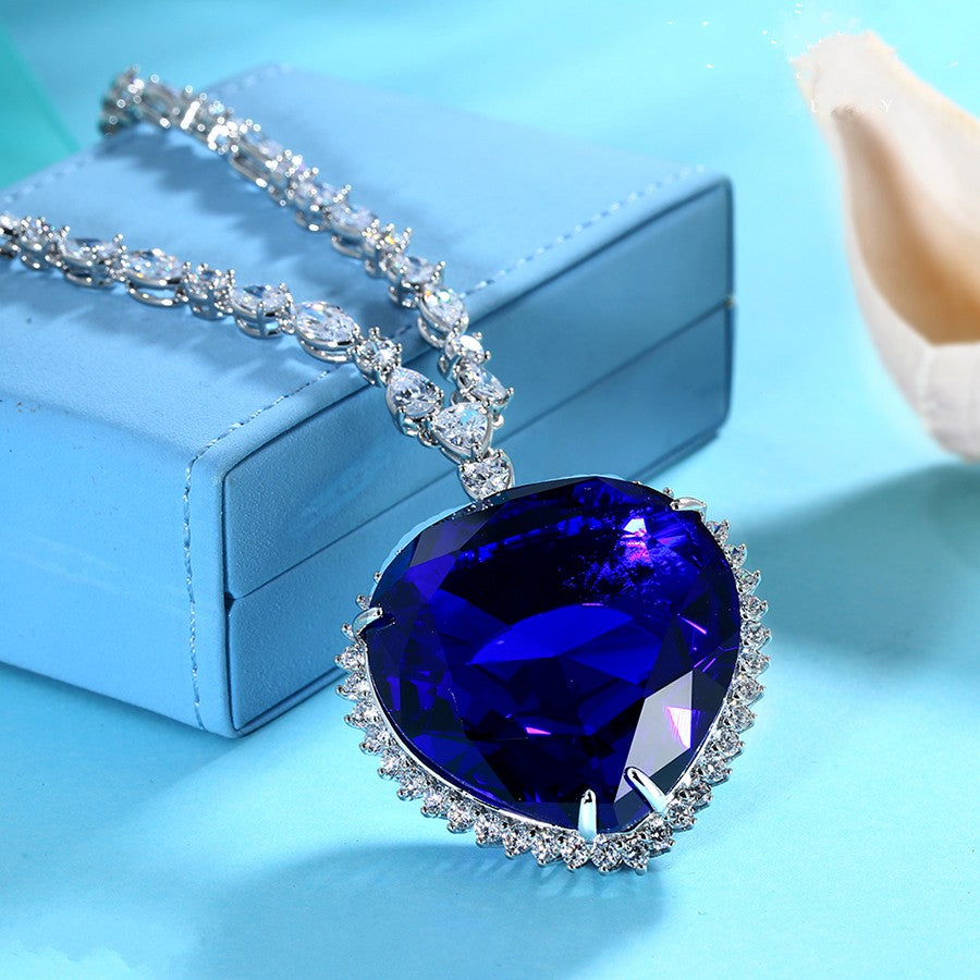 A replica of the necklace from the movie Titanic, the necklace is called the Heart of the Ocean. This is a closeup of the main pendant which shows the blue gem. Some links of the chain can also be seen. This necklace is sitting on a blue jewelry box and the pendant is hanging down off the box. This is on a blue background.